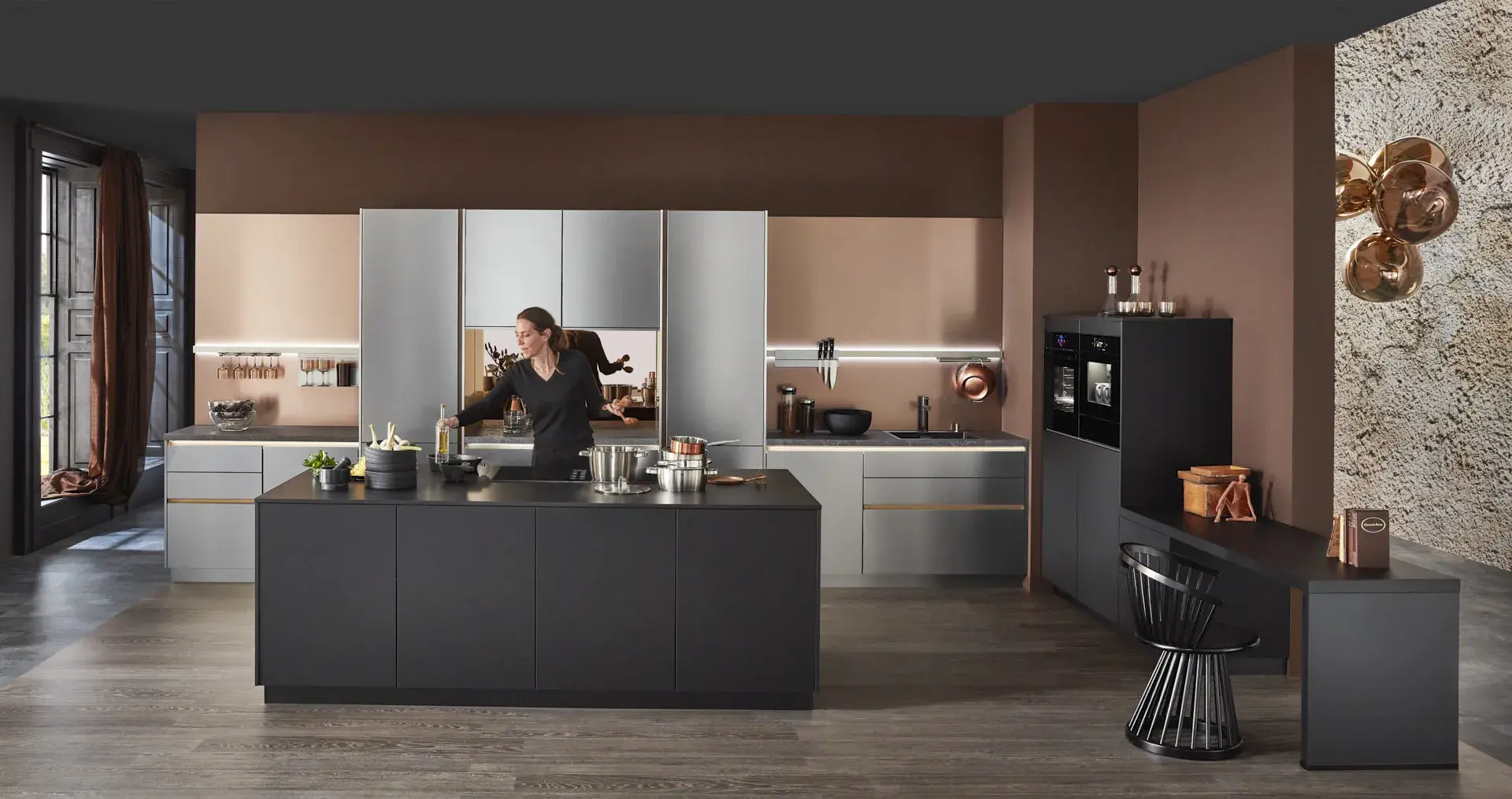 Hauptschuss, Mix aus Keramik und Bronzenachbildung, Kücheninsel mit Bosch Induktionskochfeld und Muldenlüfter, schwarzer Schrank, Edelstahlfronten, Bronze Nischenrückwand, 2 Bosch Einbaubacköfen, Messing Spüle, Model ist hinter Kücheninsel